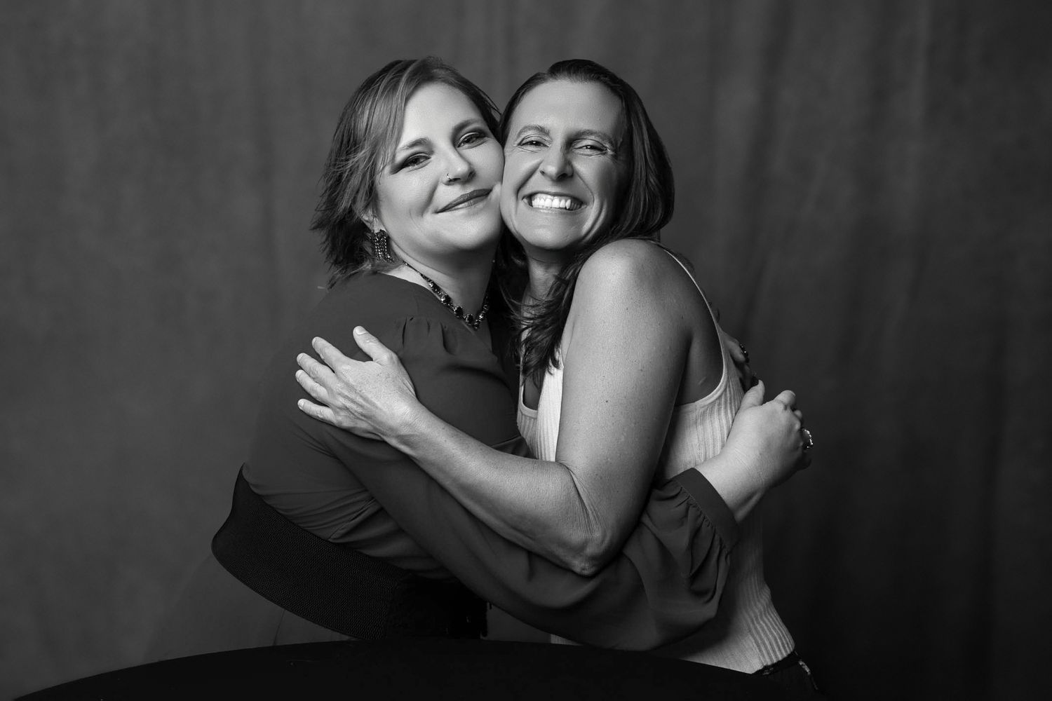 Two women share a warm embrace, smiling joyfully in a black and white portrait photo.