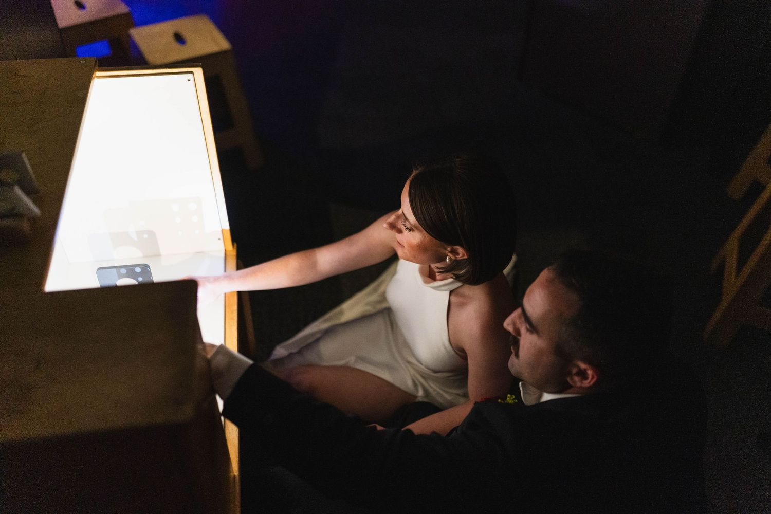 Couple sharing an intimate moment under warm lamp light in a dark room, bride in white dress and groom in suit.