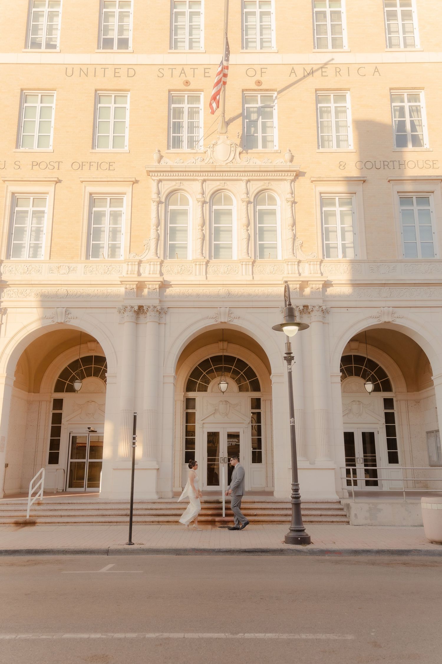 Couple poses in front of a grand U.S. Post Office and Courthouse building with arched entrances and American flag.