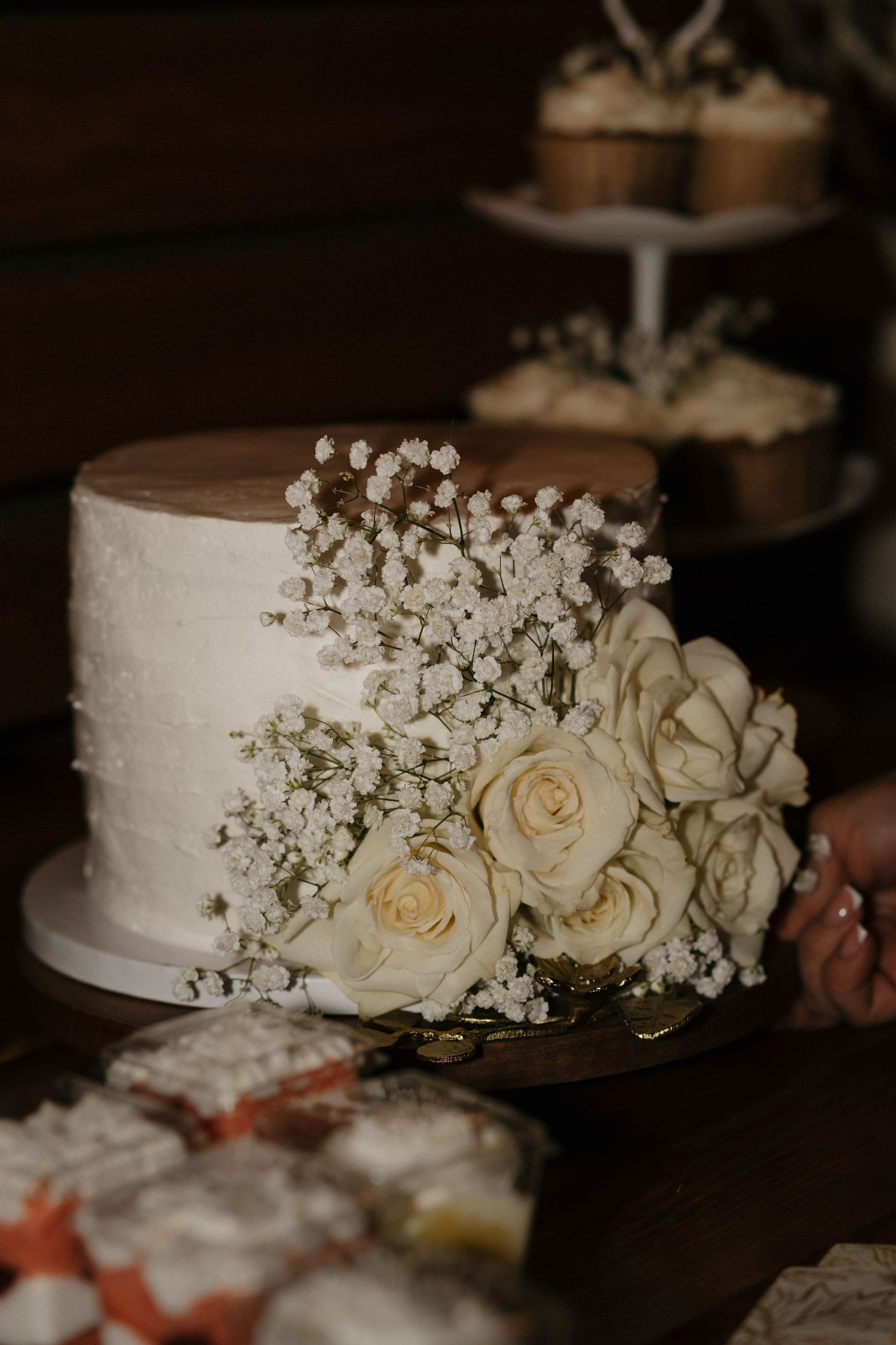White wedding cake adorned with cream roses and baby's breath flowers on a wooden cake stand at a reception.
