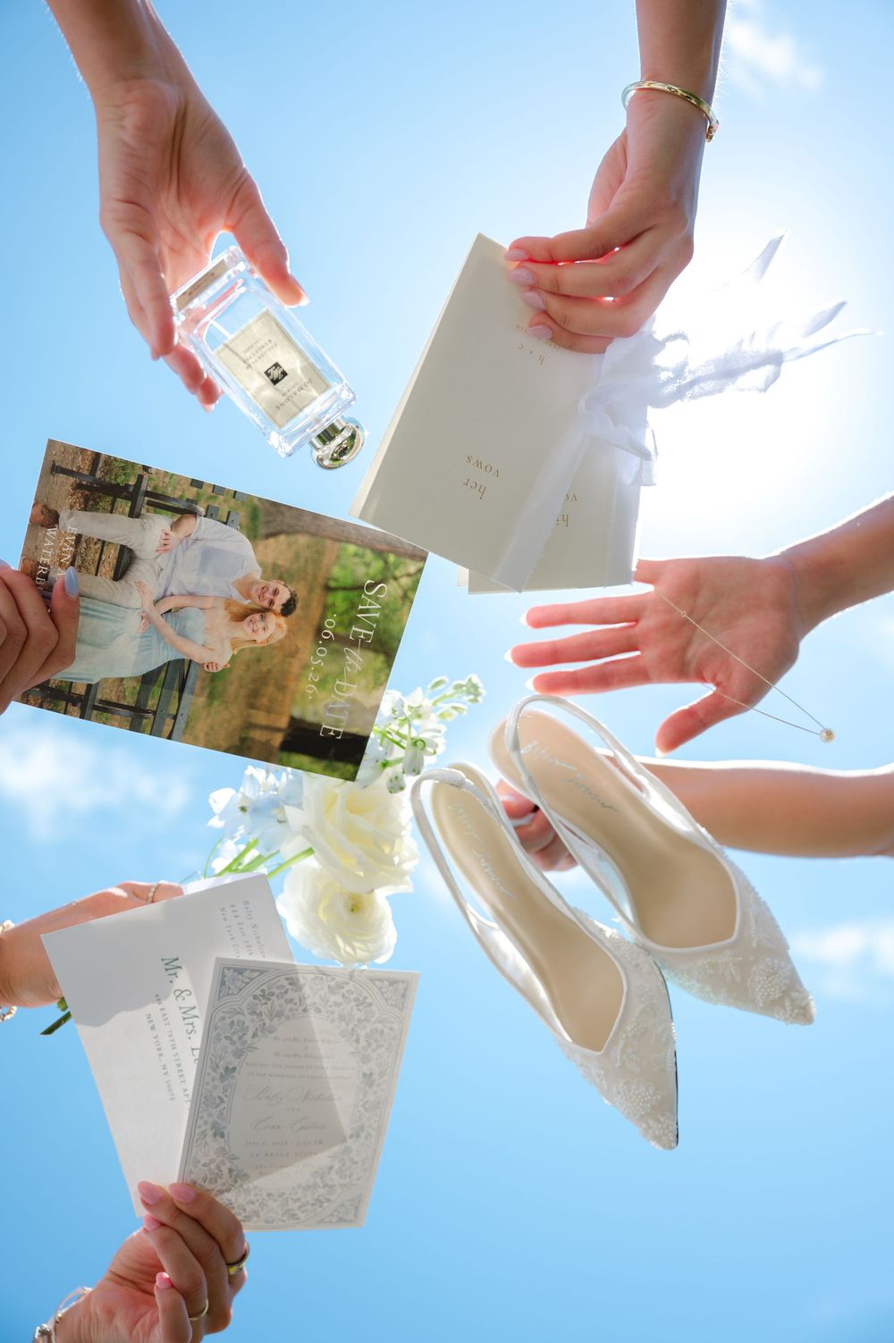 Hands holding wedding items including shoes, invitation, perfume, and photo against blue sky.