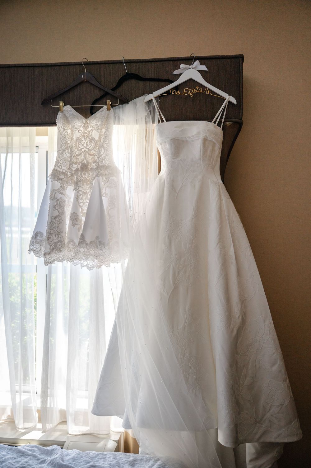 Two white wedding dresses hanging on black hangers in front of a sunlit window, including a lace gown and a satin A-line dress.