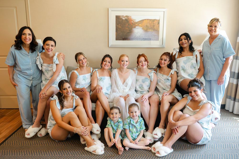 Bride in white robe poses with bridesmaids in matching blue floral pajamas and two young children in a hotel room.