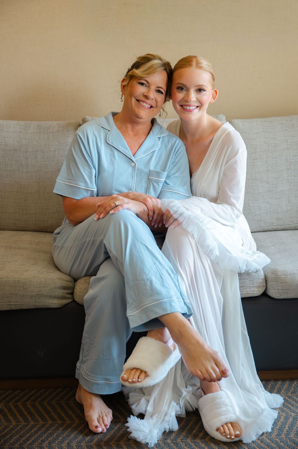 Bride in white robe and mother in blue pajamas sitting together on a couch smiling before the wedding.