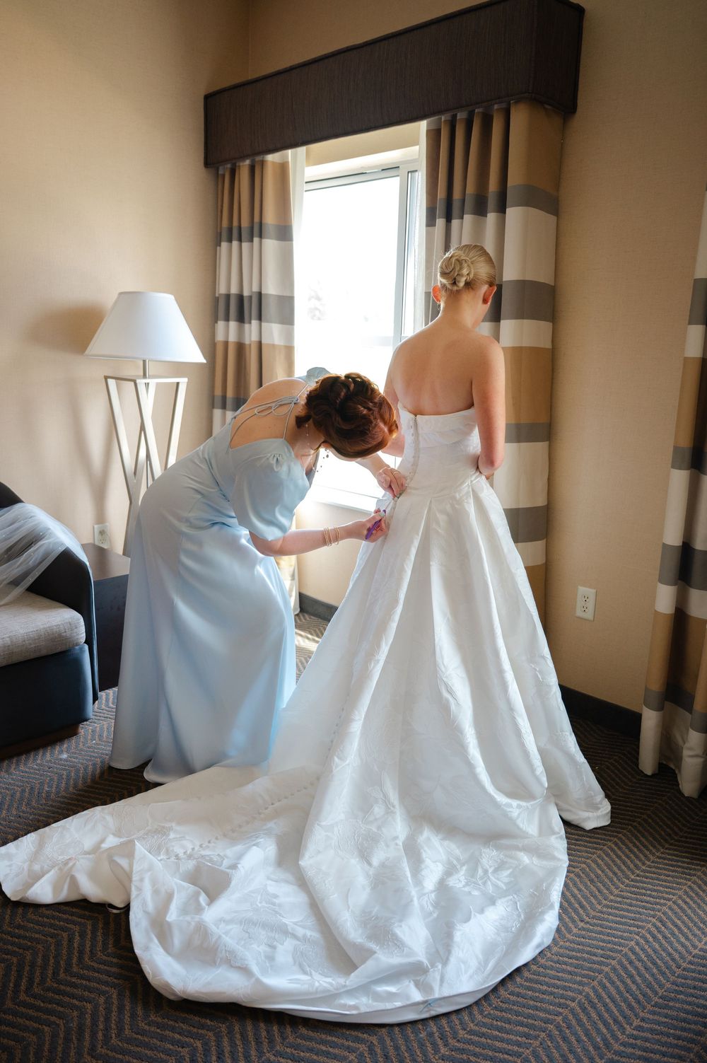Bridesmaid helping bride button up her white strapless wedding gown in a hotel room before the ceremony.