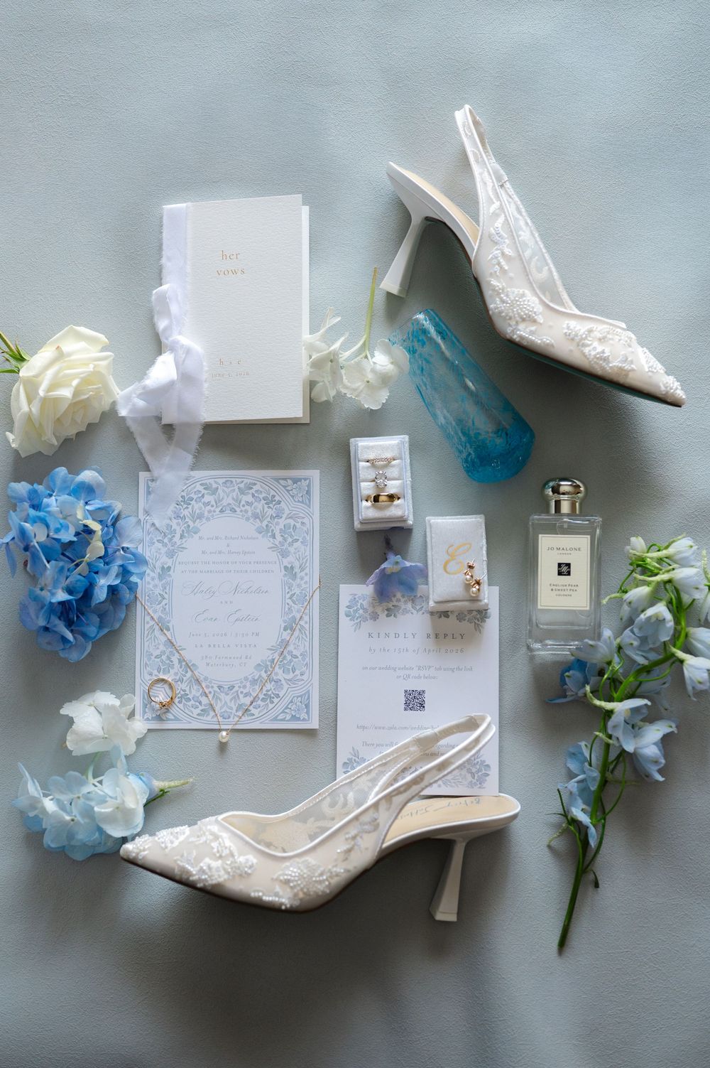 Elegant wedding flat lay with lace heels, blue hydrangeas, Jo Malone perfume, invitation suite, and gold rings on a soft blue background.