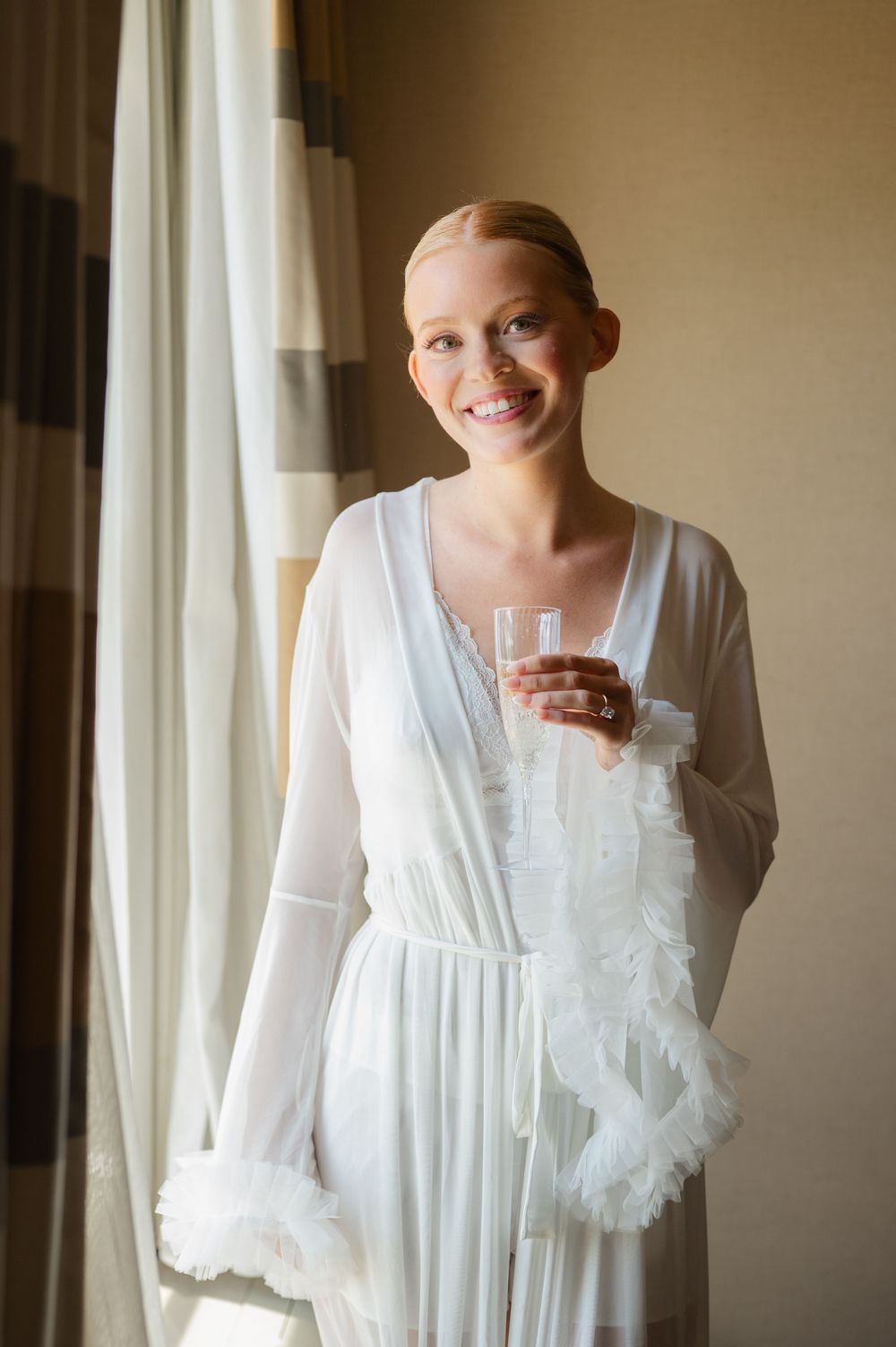 Smiling bride in white sheer robe holding a glass near a window on her wedding day.
