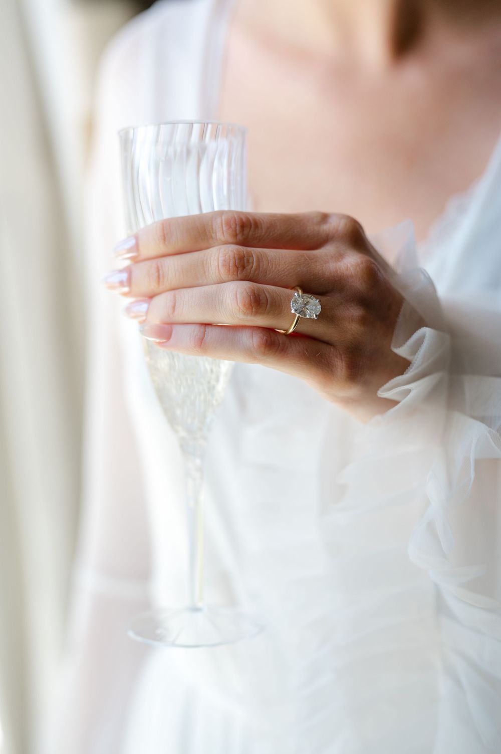 Bride holding a champagne flute with a sparkling diamond engagement ring on her finger, wearing a white wedding gown.