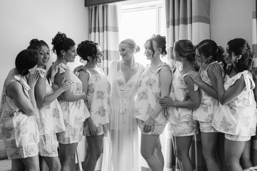 Bride in white robe surrounded by bridesmaids in floral rompers during bridal preparations, black and white photo.