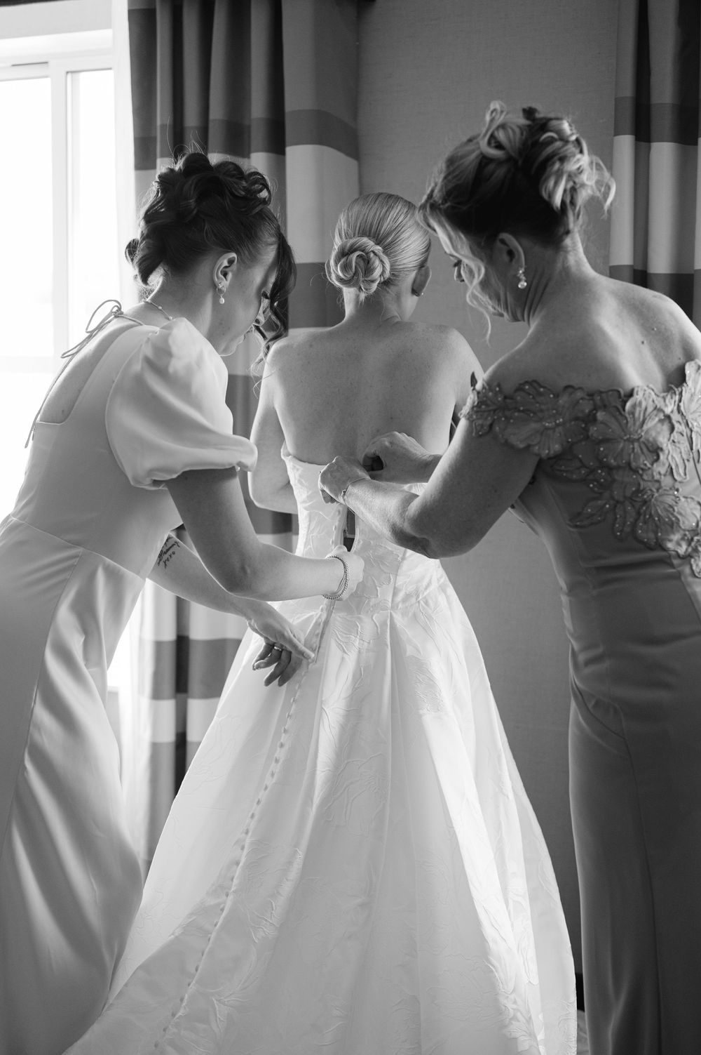 Black and white photo of a bride being helped into her wedding gown by two bridesmaids in elegant dresses.