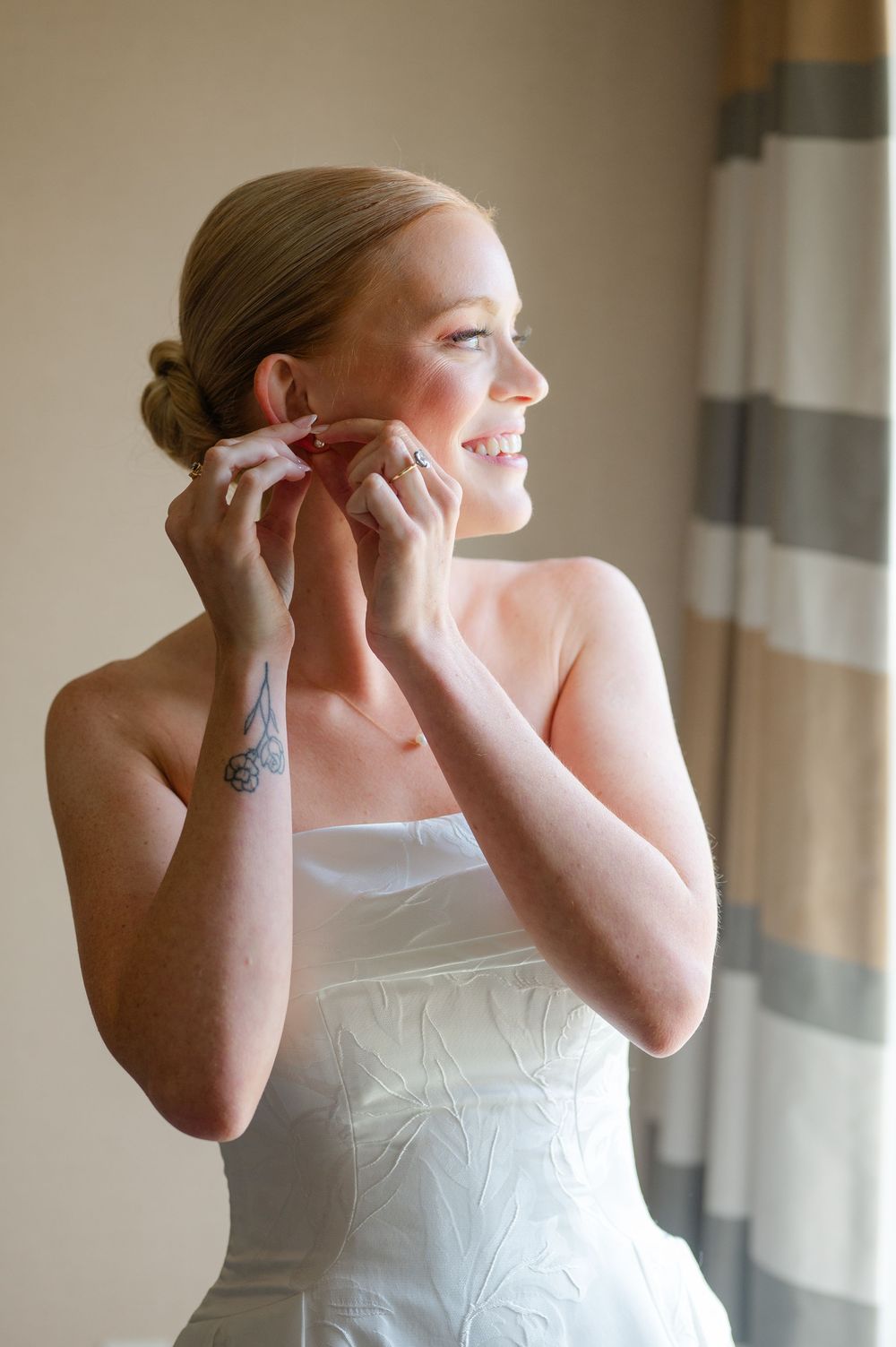 Smiling blonde bride in a strapless white wedding gown putting on earrings while getting ready on her wedding day.