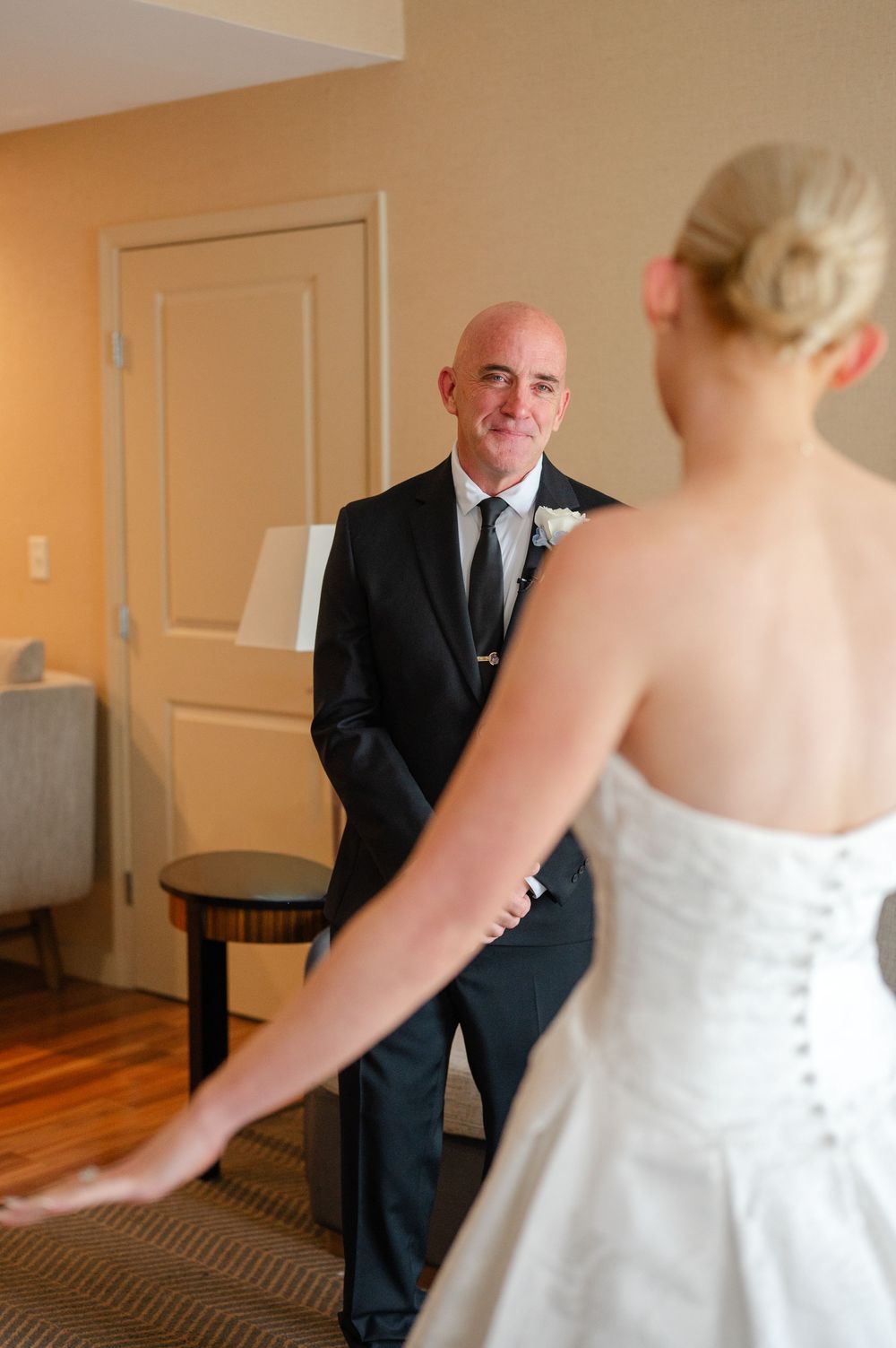 A bald groom in a black suit smiles at his bride in a white strapless wedding gown during their first look.