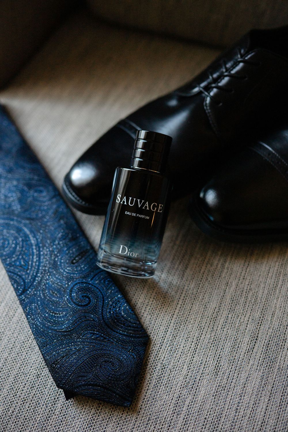 Dior Sauvage Eau de Parfum bottle beside a blue paisley tie and black leather dress shoes on a neutral surface.