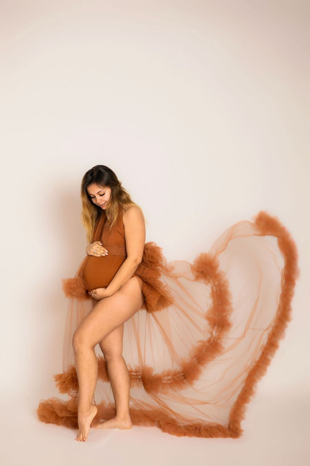 Pregnant woman in rust bodysuit with flowing tulle skirt poses gracefully in a studio maternity photoshoot.