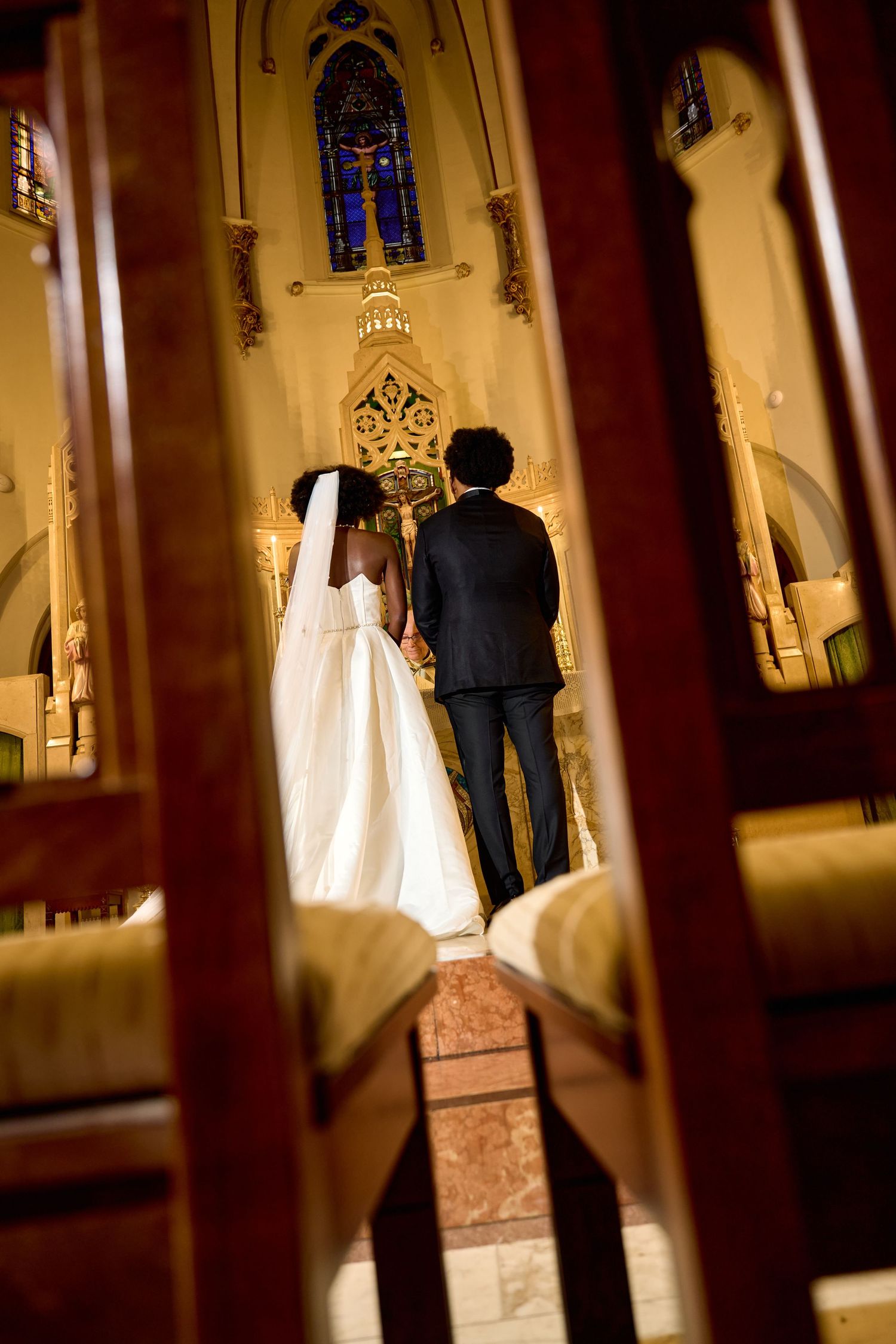 A bride and groom stand at the altar of an ornate church during their wedding ceremony, viewed through wooden pews.
