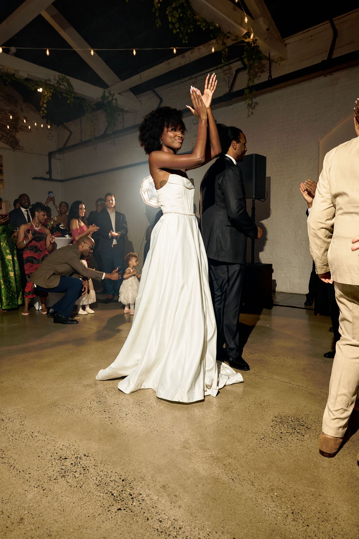 Bride in white gown and groom share first dance while wedding guests clap and celebrate in warmly lit venue.