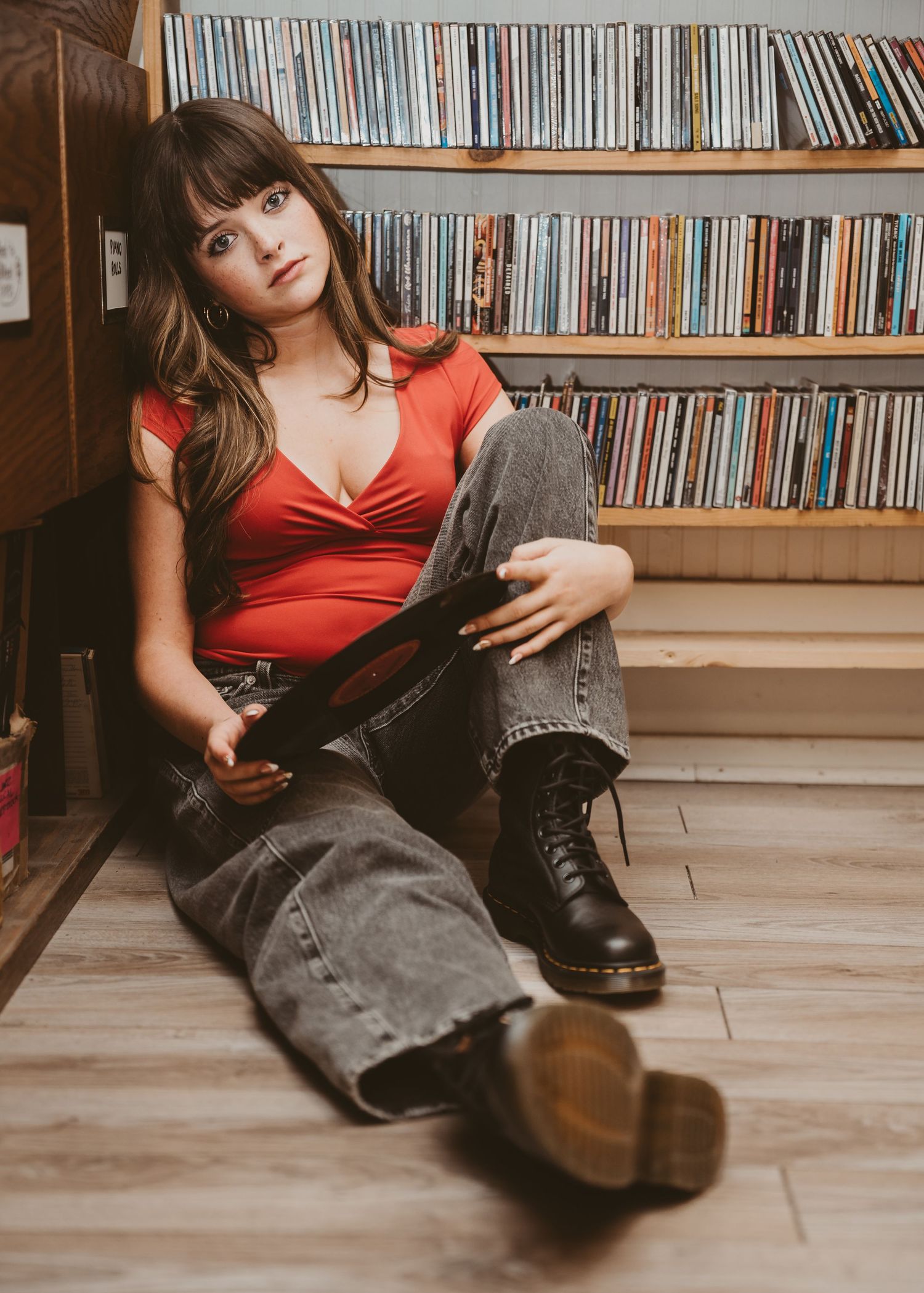 Young woman in red top and Dr. Martens sits on floor of vinyl record shop holding an album.