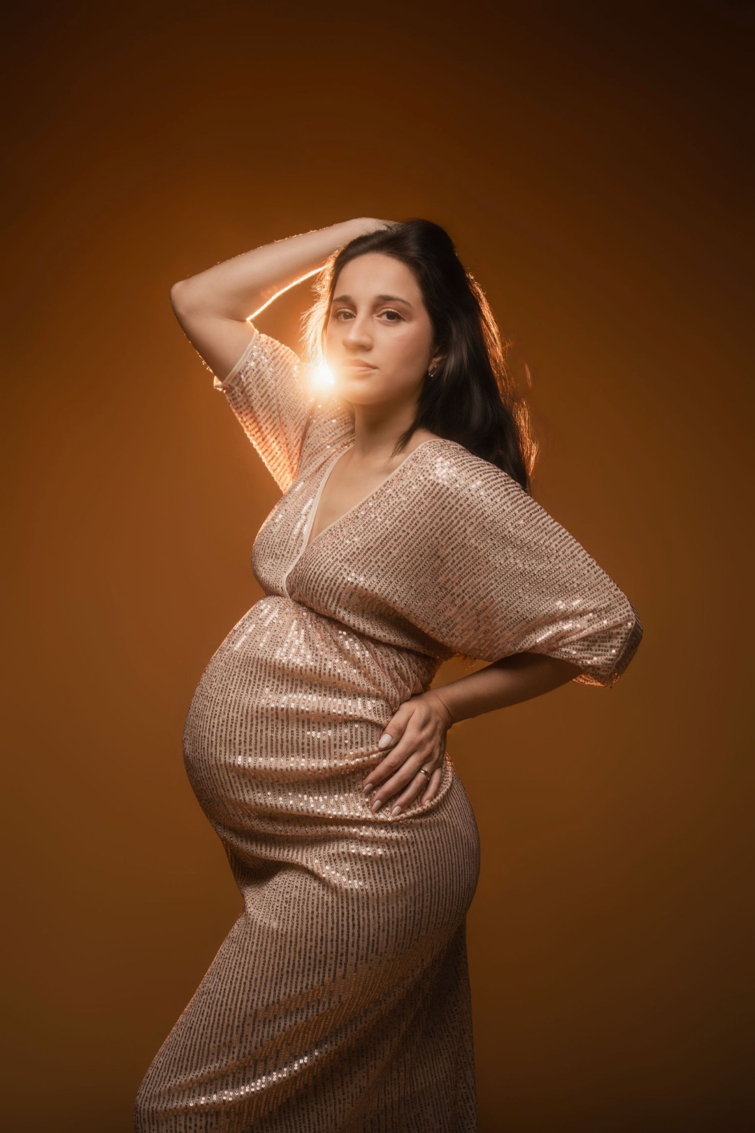 Pregnant woman in gold sequin dress posing elegantly with backlit glow on warm brown background.