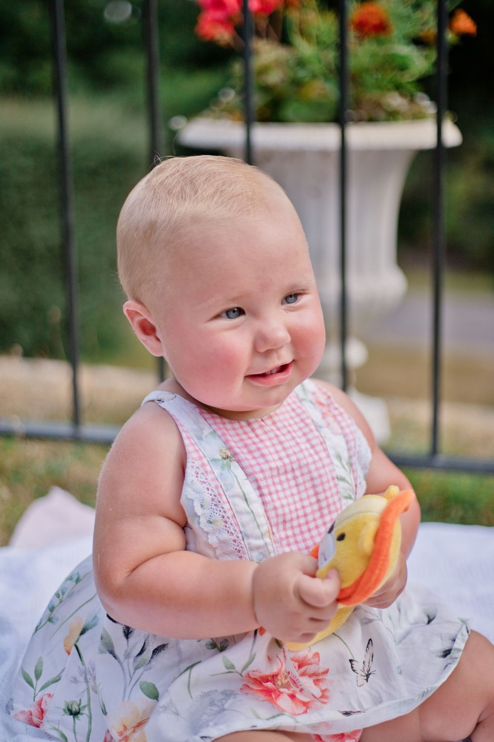 Smiling baby girl in floral dress holding colorful toy outdoors near iron fence with flowers.