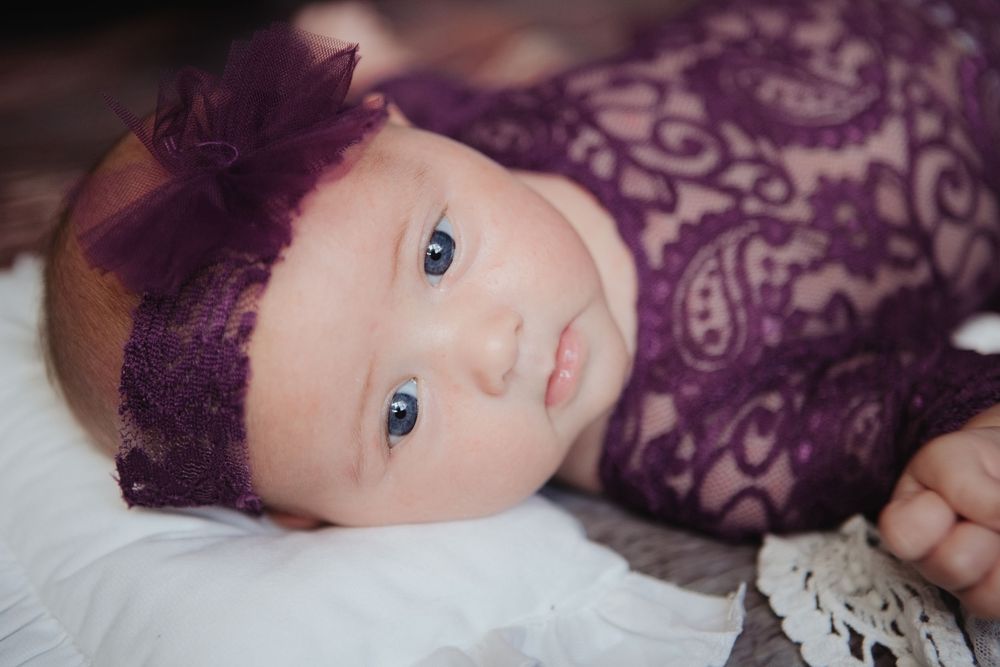 Newborn baby girl in purple lace wrap and floral headband lying on white blanket.