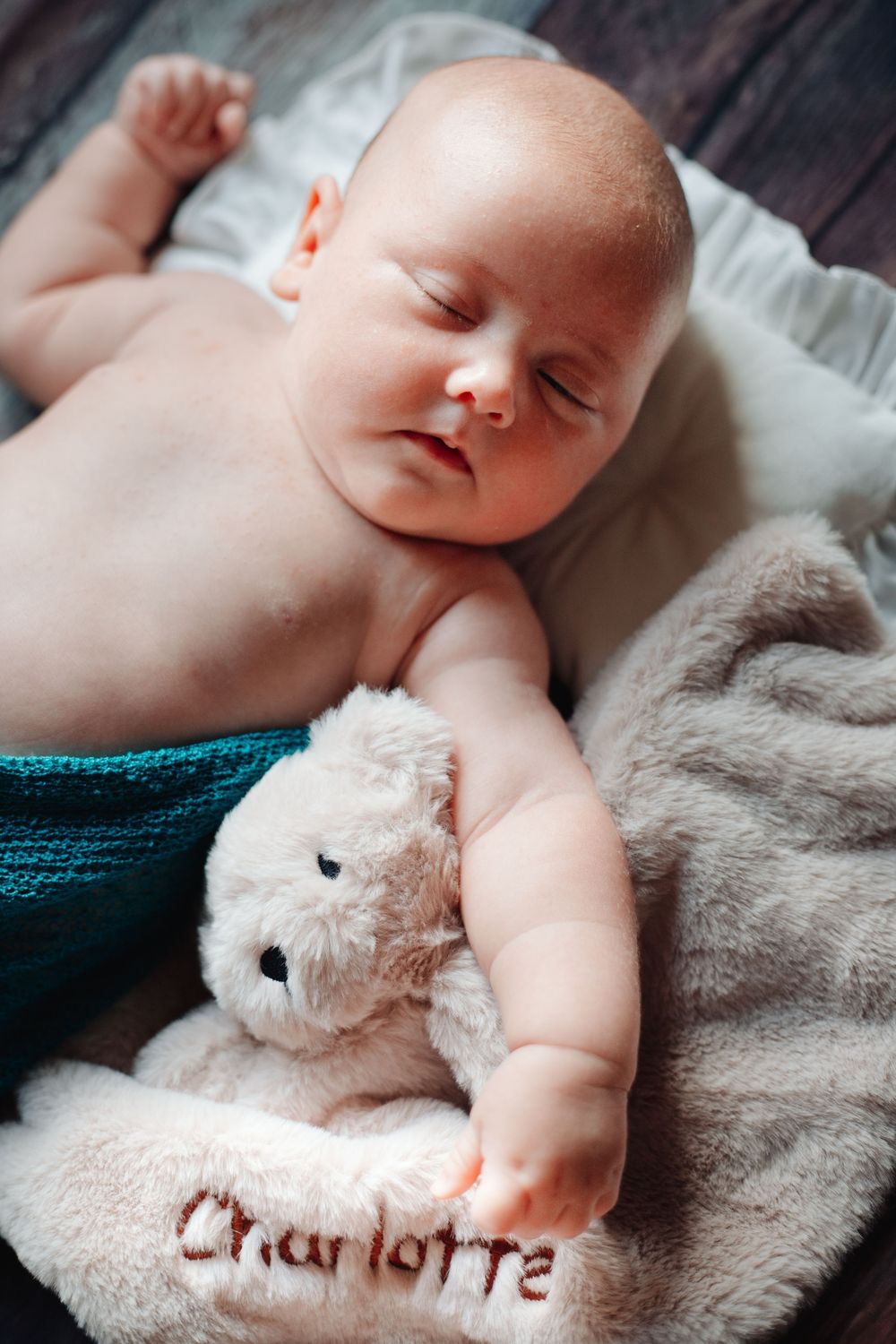 Sleeping newborn baby in teal wrap beside personalized Charlotte teddy bear on wooden floor.