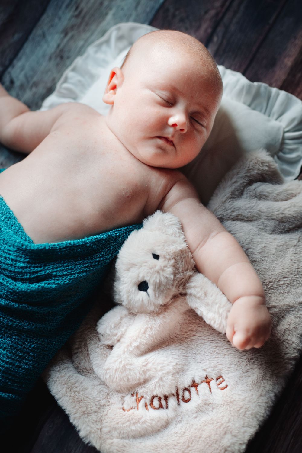 Peaceful newborn baby sleeping next to embroidered Charlotte teddy bear on rustic wooden surface.