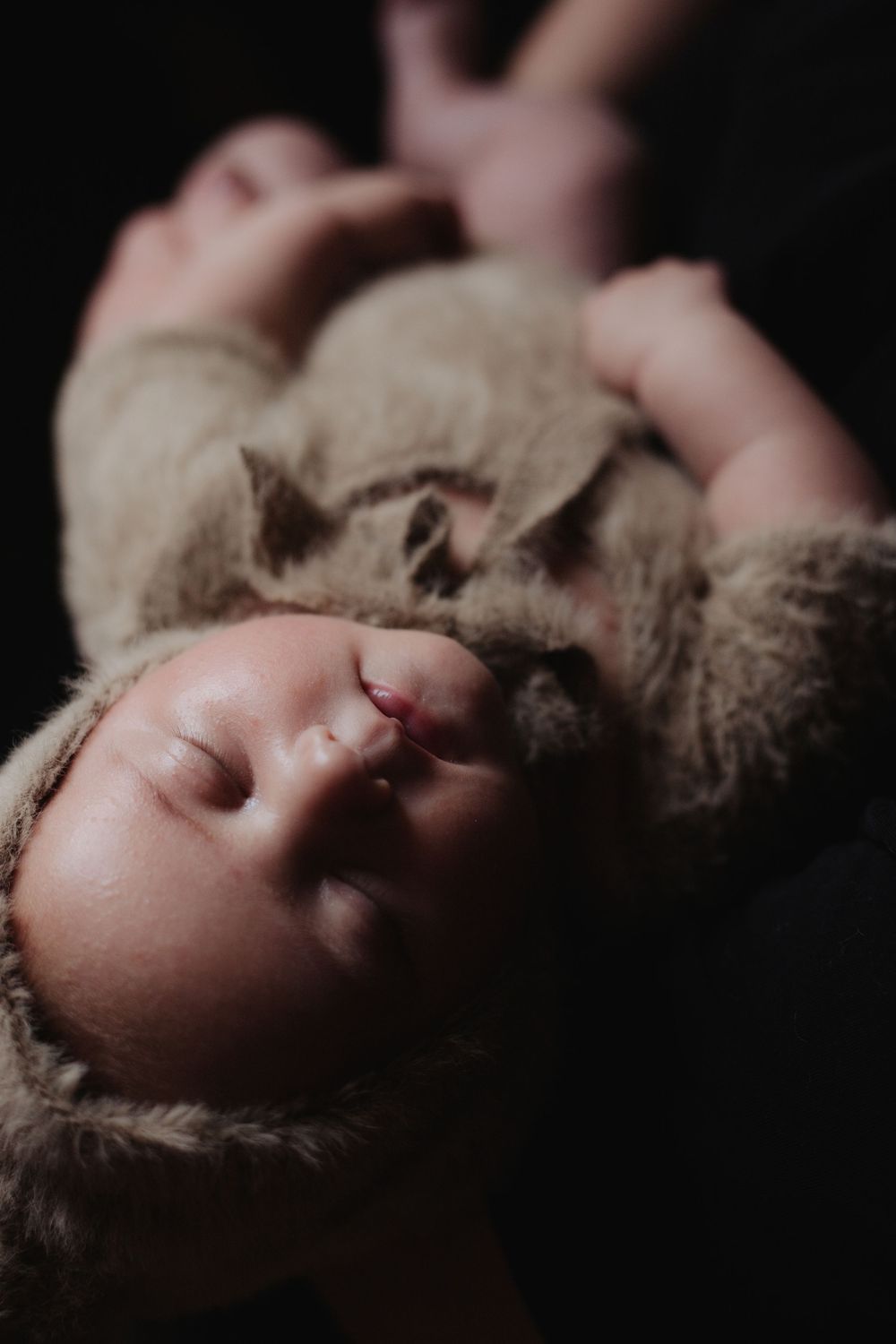 Sleeping newborn baby wrapped in soft brown fuzzy bear outfit in dark moody setting.