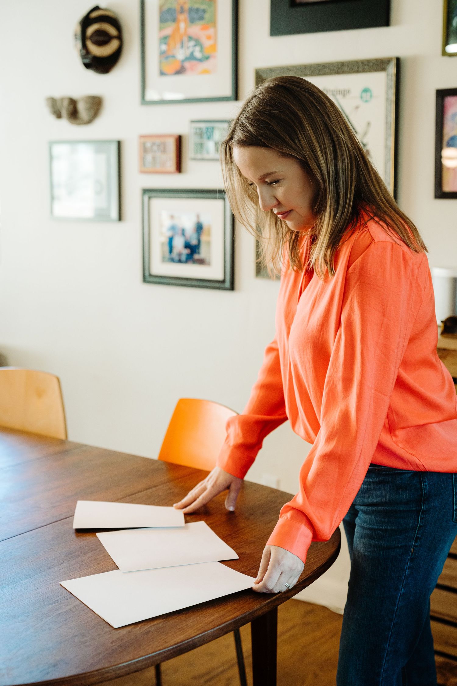 Woman in orange top arranging white papers on a wooden dining table with gallery wall art in background.
