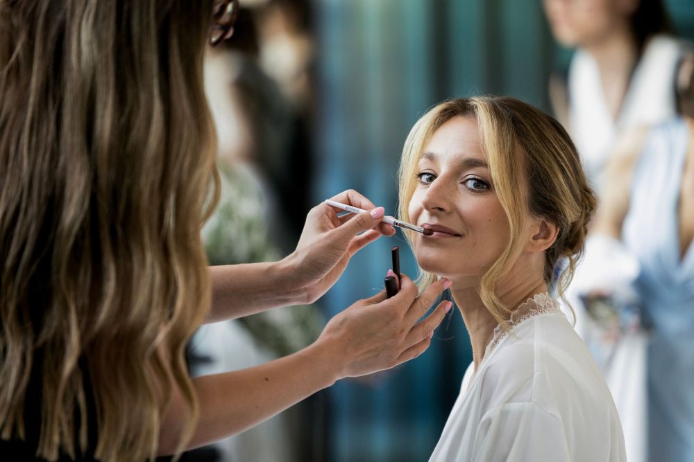 Professional makeup artist applying lipstick to a smiling blonde bride in a white robe during wedding preparation.