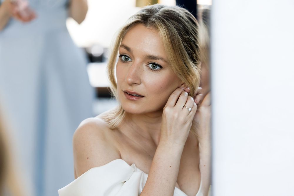 Elegant blonde woman in off-shoulder white top touching her ear, wearing a delicate ring, soft natural makeup.