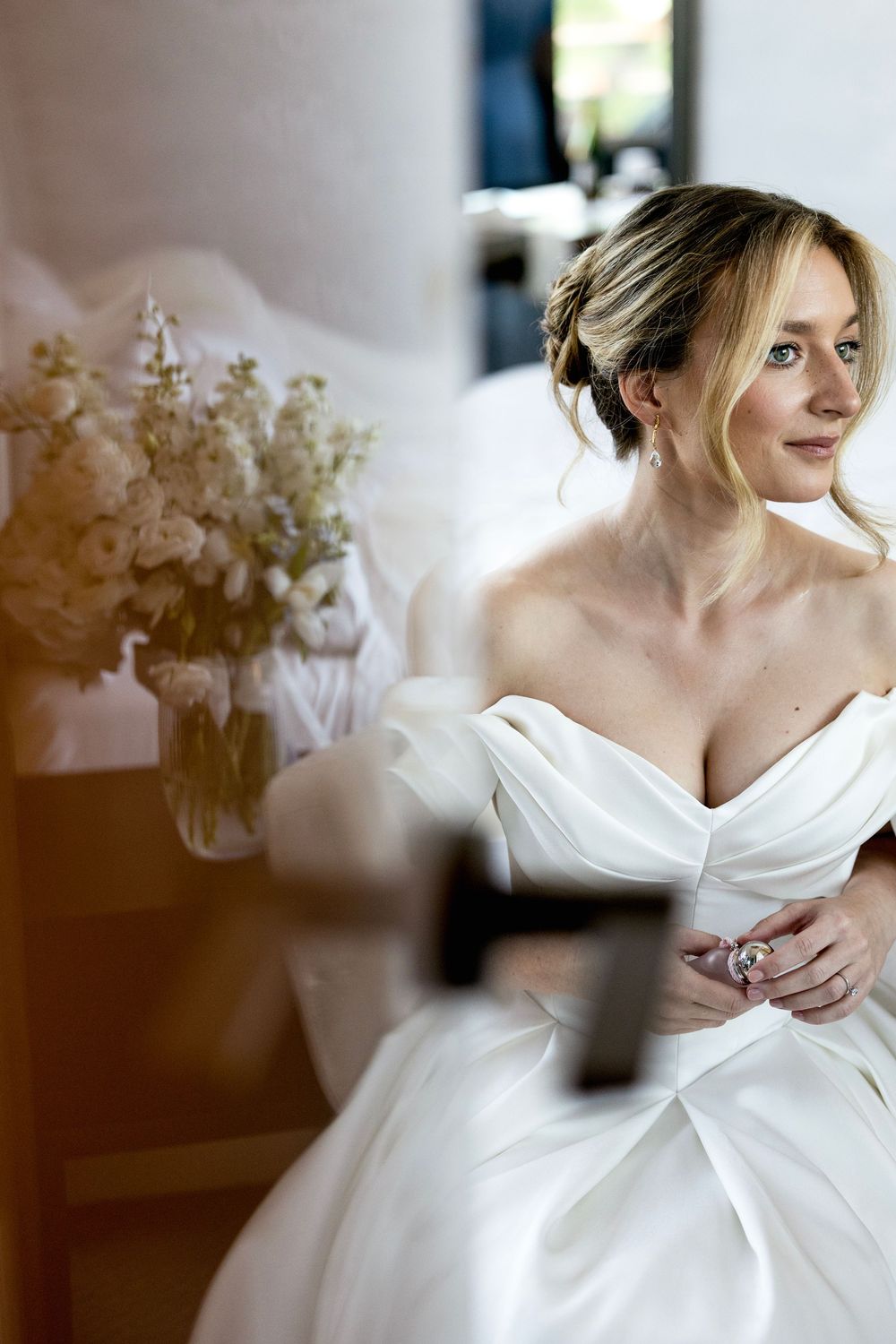 Elegant bride in off-shoulder white ballgown sits gracefully, wearing diamond ring, with floral bouquet nearby.