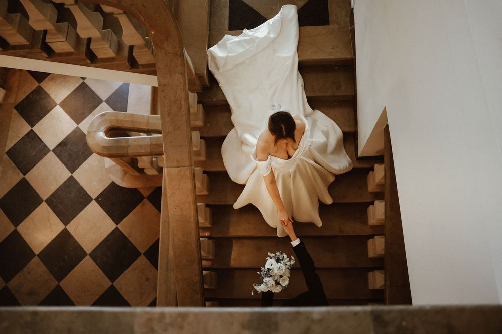 Bride in wedding dress sitting on wooden staircase holding bouquet, aerial view with checkered floor below.