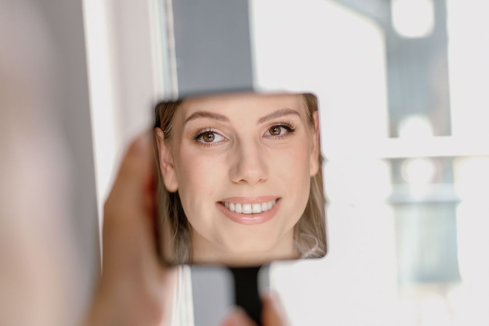 Woman smiling at her reflection in a handheld mirror, showcasing healthy skin and bright teeth.