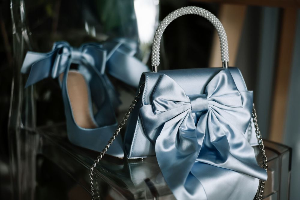 Light blue satin handbag with large bow and pearl handle displayed alongside matching blue heels.