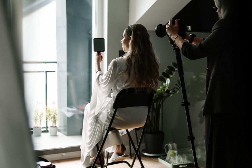 A woman in a white robe sits by a window holding a mirror while a photographer captures her portrait indoors.