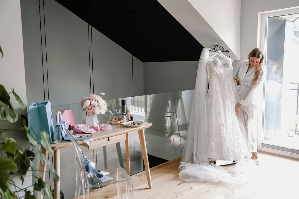 Bride in white robe admiring wedding gown in bright room with floral arrangements and accessories on desk nearby.