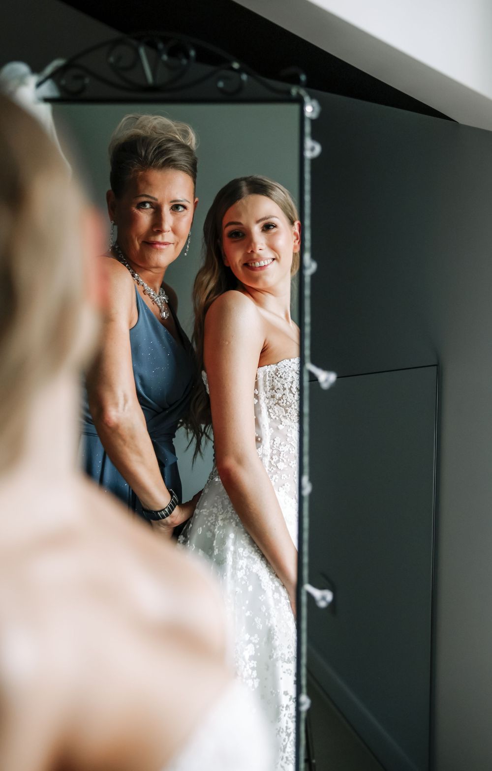 Bride in white lace gown with woman in blue dress smiling together in mirror reflection.