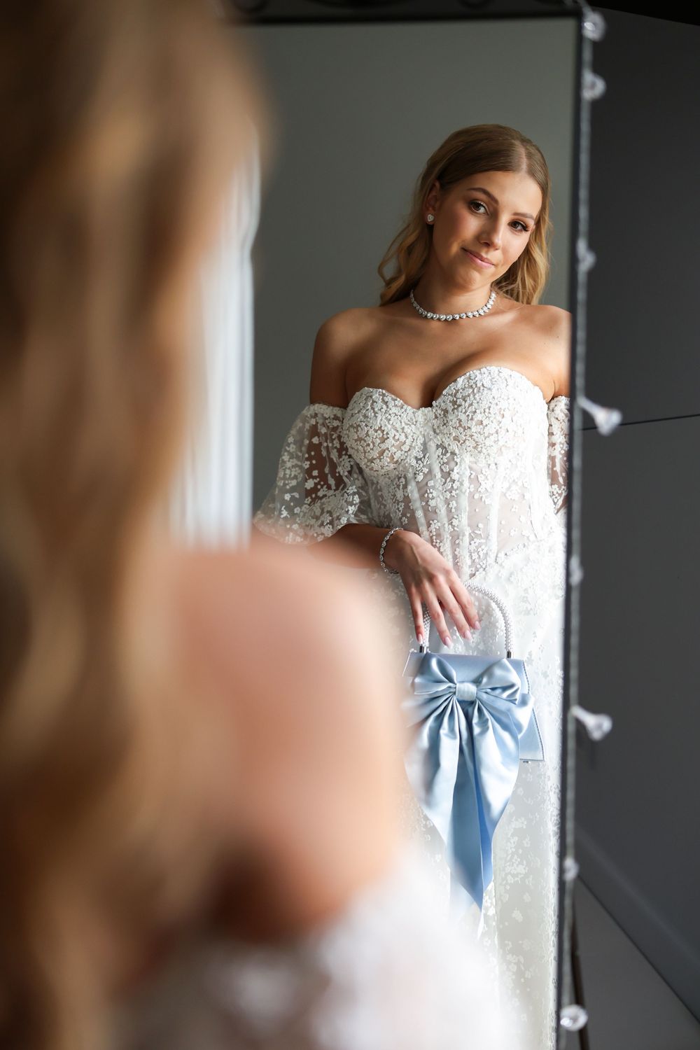 Bride in white lace off-shoulder gown holding blue bow purse, admiring her reflection in a Hollywood mirror.