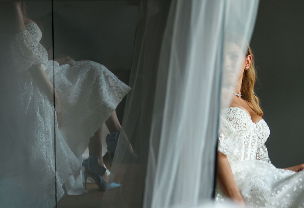 Bride in lace wedding dress sits by sheer curtains, reflection visible in mirror showing heeled shoes.