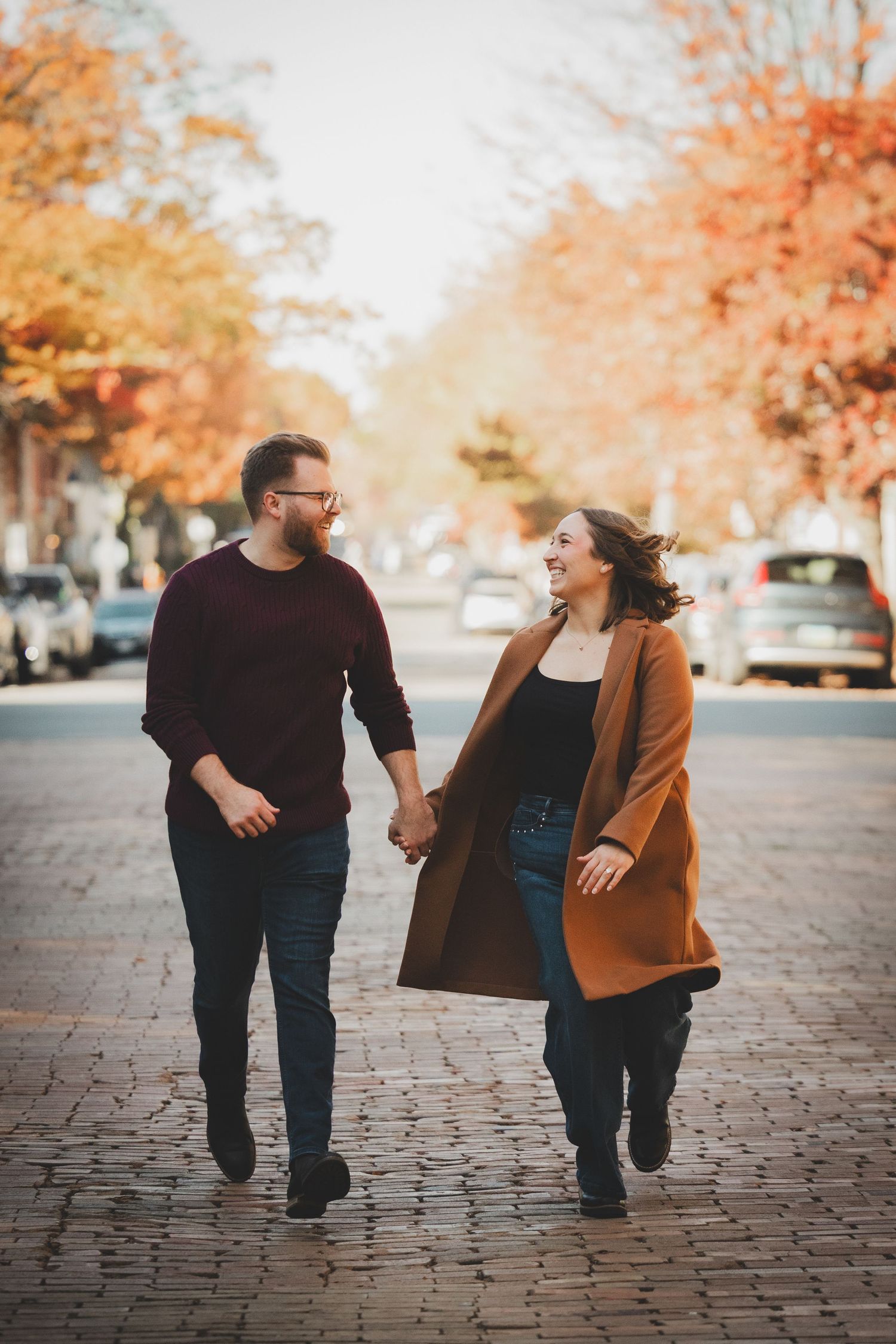 Couple walking hand in hand and smiling at each other on a cobblestone street during their Old Town Alexandria engagement session