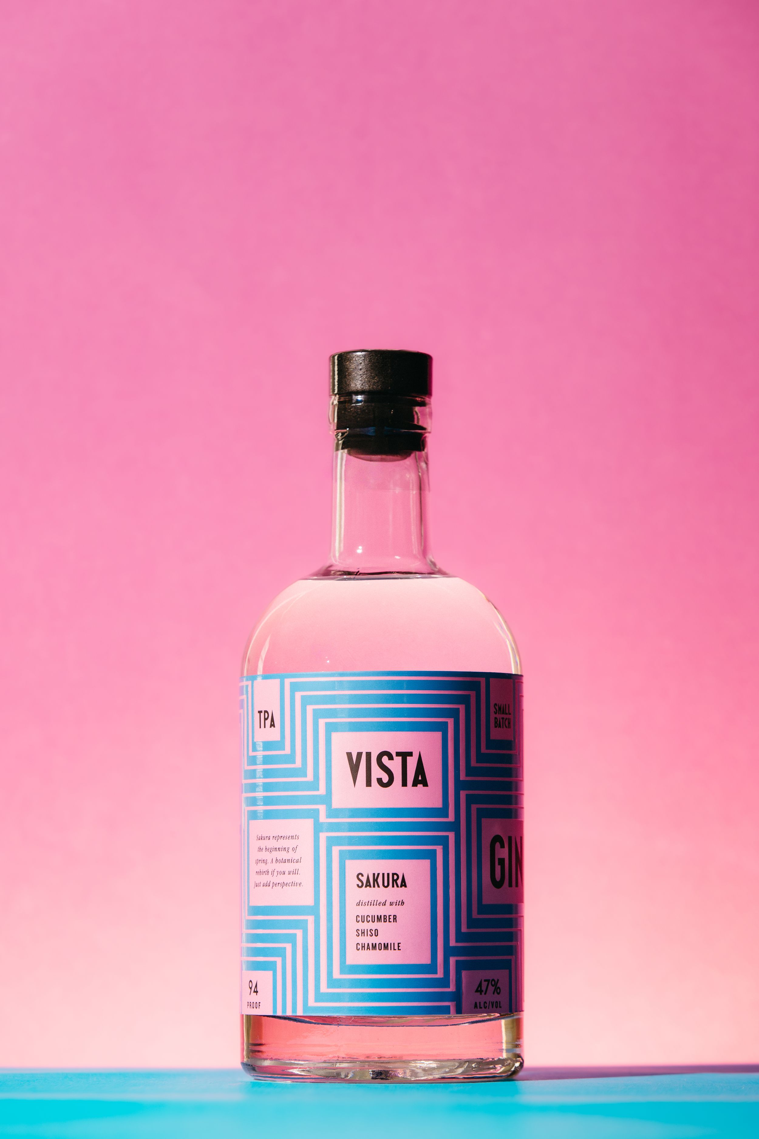 Vista Sakura Gin bottle with blue geometric label, pink liquid, against vibrant pink background on teal surface.