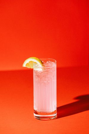 A tall glass of pink lemonade with ice and a lemon slice garnish on a vibrant red background.