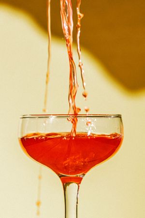 Red cocktail being poured into a coupe glass with dramatic splash against a yellow background.