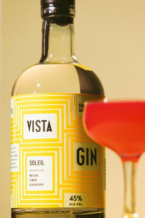 Vista Soleil Gin bottle with yellow geometric label, 45% ABV, distilled with matcha, lemon, and blackberry, beside a red cocktail.