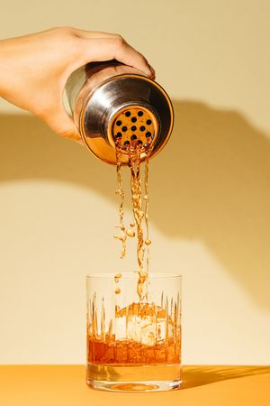 Bartender pouring amber cocktail from silver shaker into crystal glass with ice on yellow background.