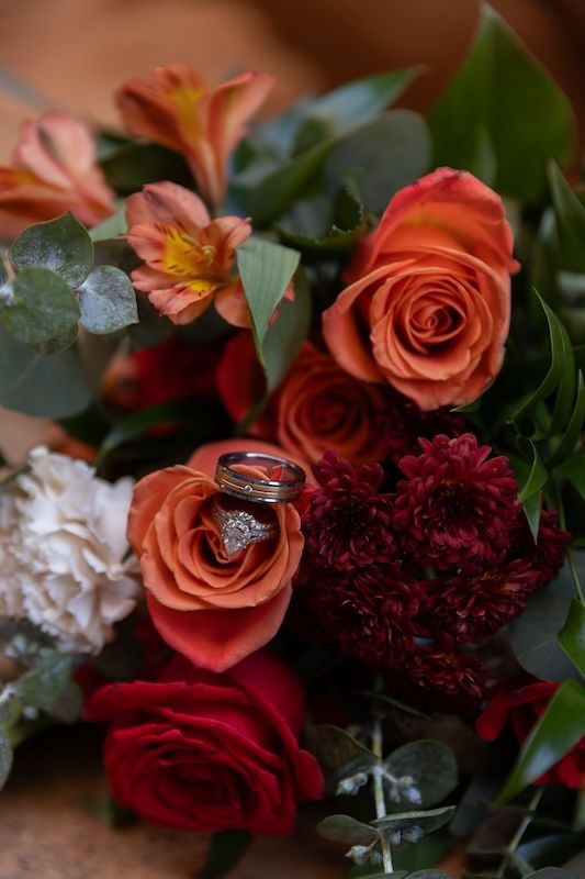 Wedding rings rest on orange and red roses in a lush bridal bouquet with burgundy dahlias and eucalyptus.