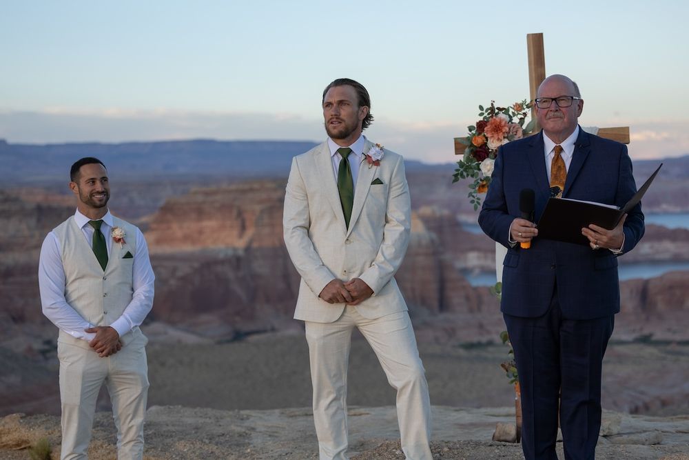 Groom in cream suit stands at outdoor canyon wedding ceremony with officiant and groomsman at sunset.