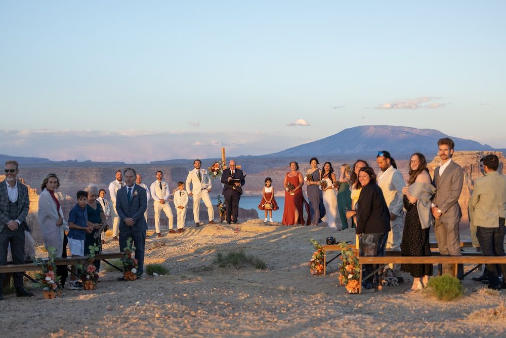 Outdoor beach wedding ceremony at sunset with guests gathered on rocky shore, mountains and water in background.