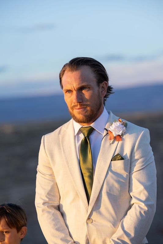 A groom in a white suit with green tie and floral boutonniere stands outdoors at a sunset wedding ceremony.