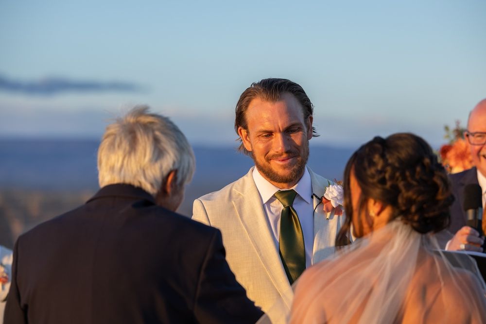 Groom in white suit smiling during outdoor wedding ceremony at sunset with ocean view.